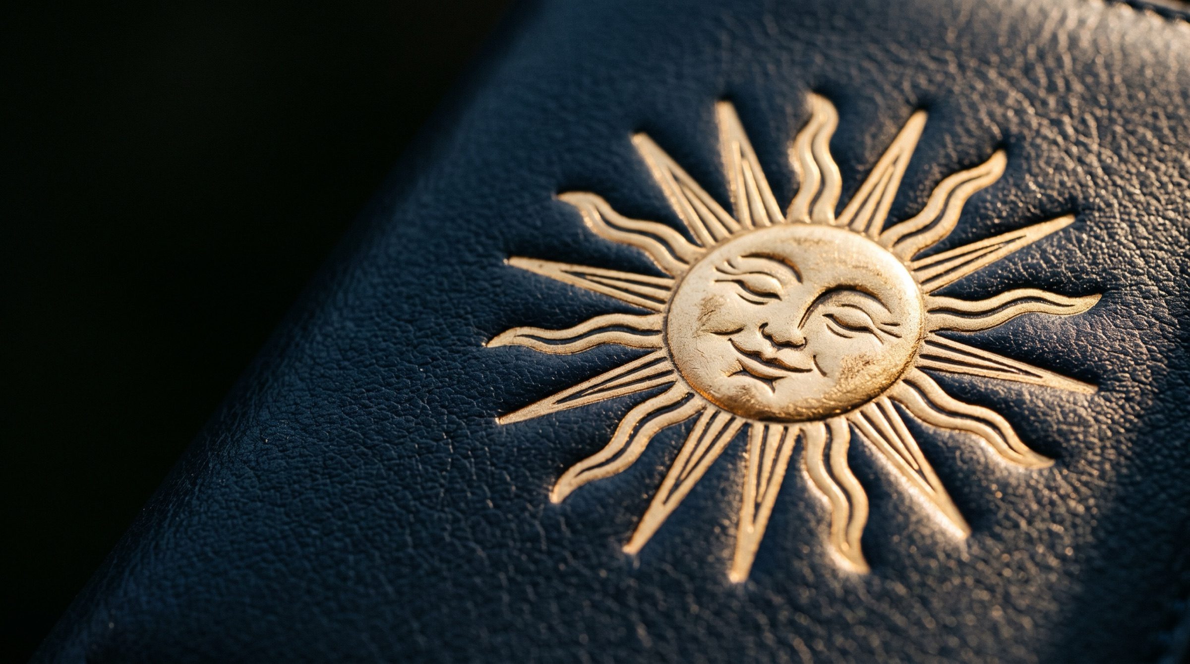 A radiant gold sun motif with alternating straight and wavy rays embossed on deep navy leather, dramatic raking light, macro detail